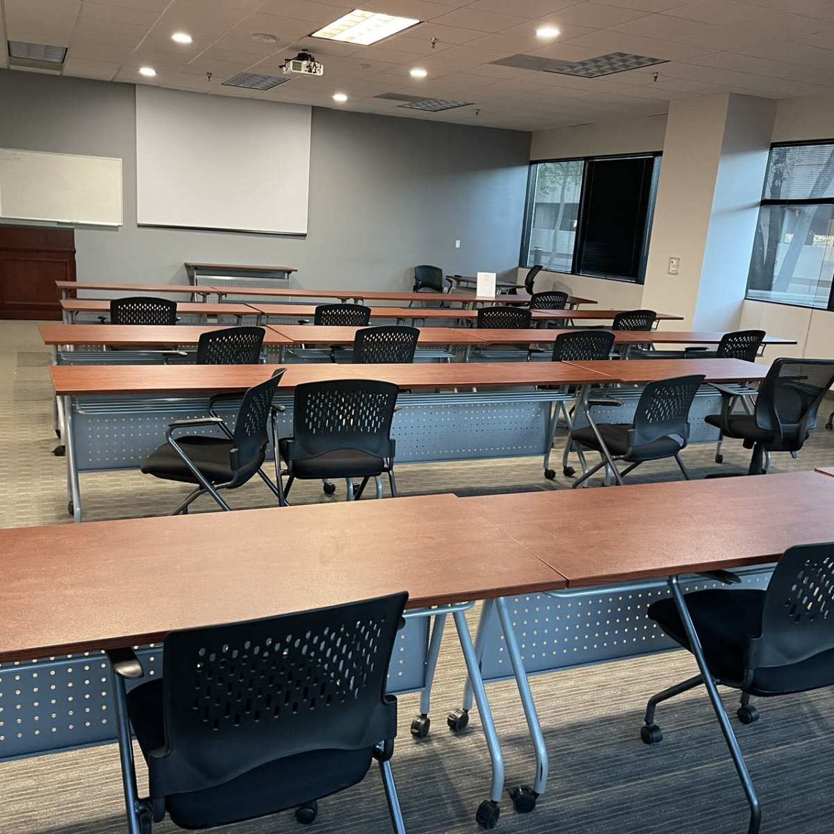 A large, empty classroom setup with multiple long tables and black chairs arranged in rows, with a projector and whiteboard at one end.