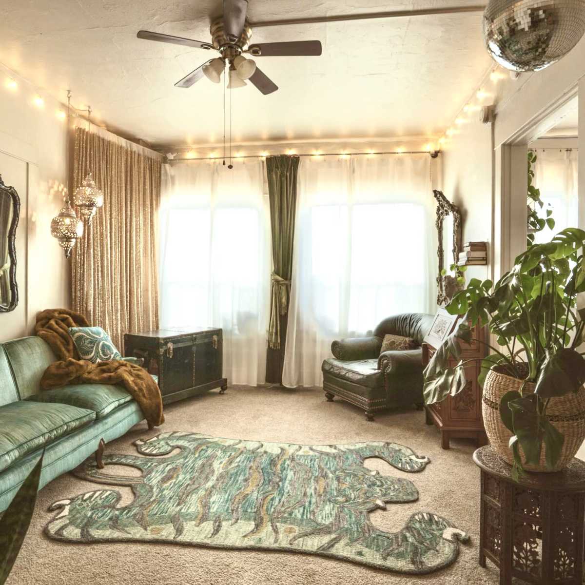 The scene features a cozy living room with a green sofa, patterned area rug, large windows, and decorative plants, illuminated by string lights.