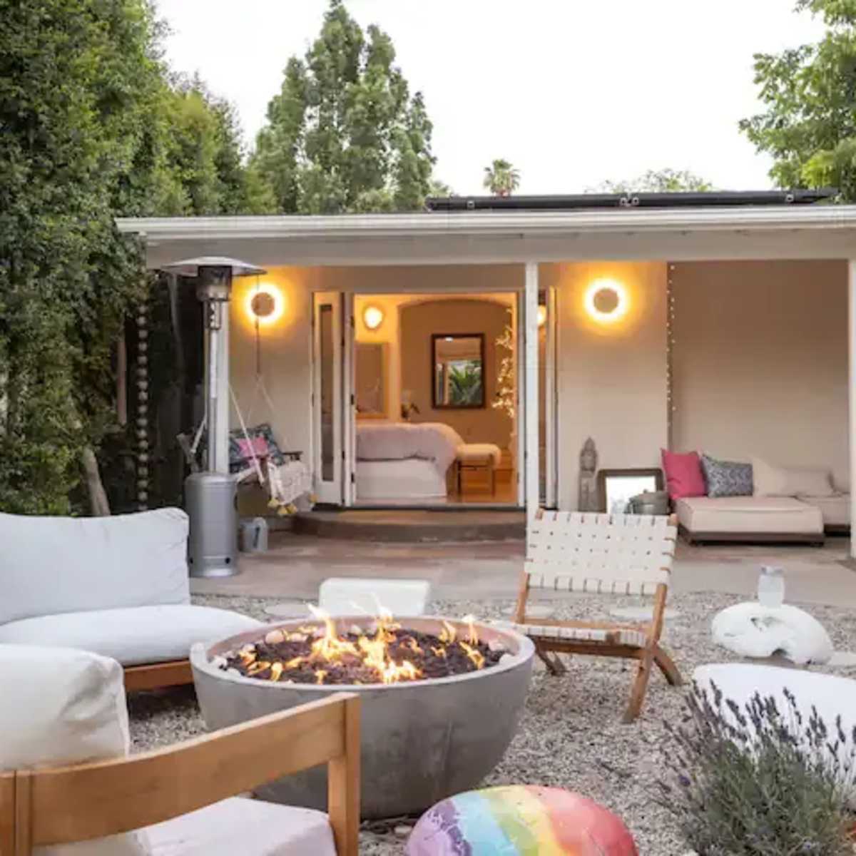 A cozy outdoor seating area features a circular fire pit surrounded by comfortable chairs and a view into a brightly lit interior space.