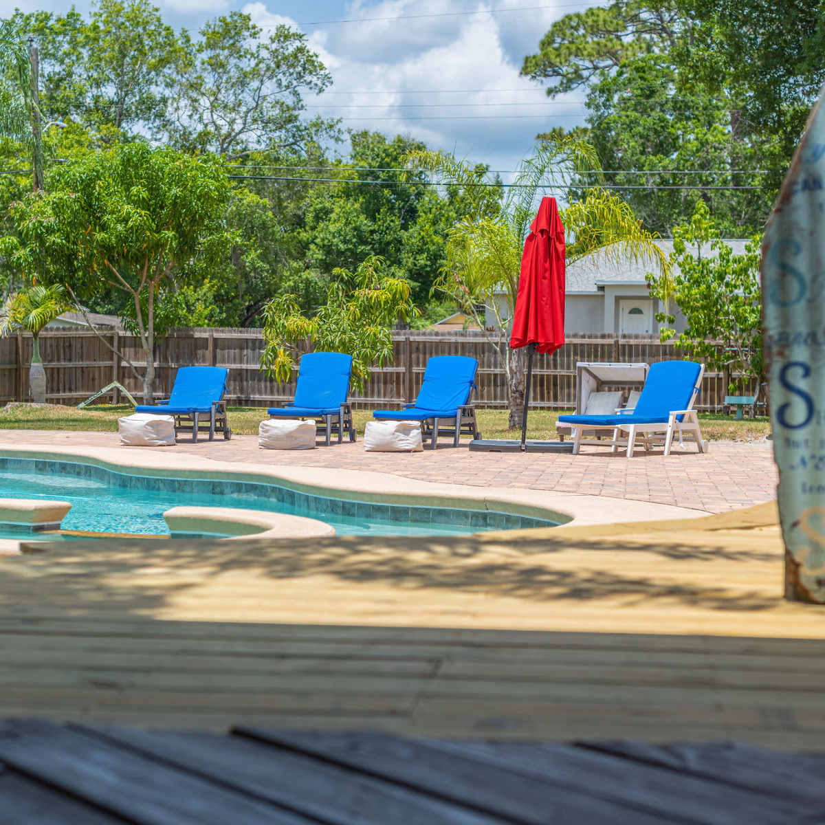A swimming pool with lounge chairs and a red umbrella is surrounded by greenery in a fenced backyard.
