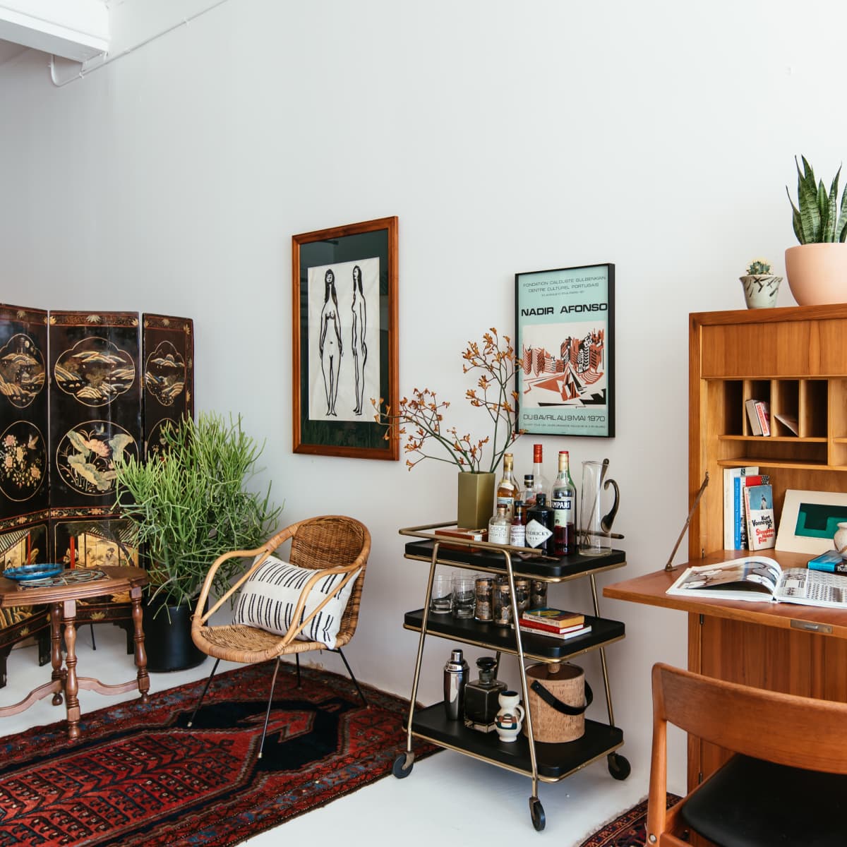 A stylish interior living space featuring a wooden desk, a bar cart, a variety of plants, and decorative wall art.