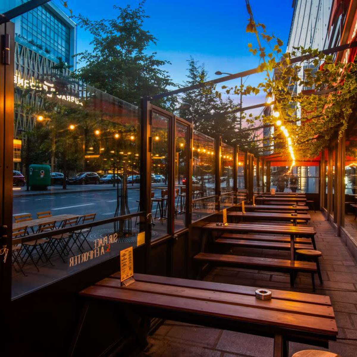 An outdoor café seating area with wooden benches lines a city street, illuminated by string lights at dusk.
