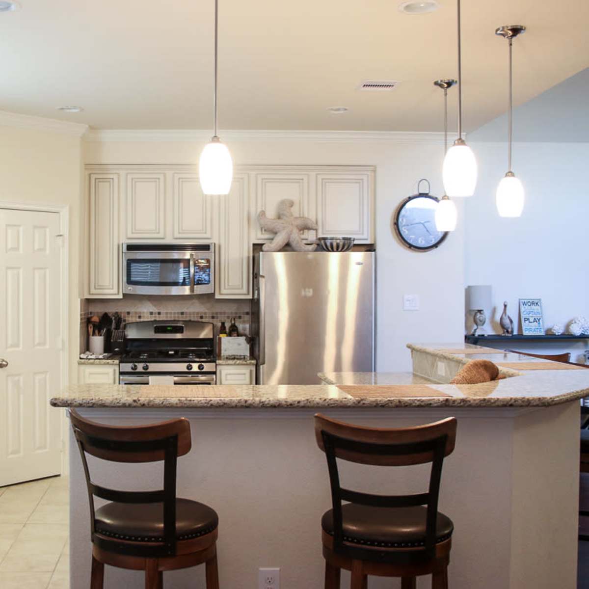 A modern kitchen with light-colored cabinetry, stainless steel appliances, and a granite countertop bar with two stools.