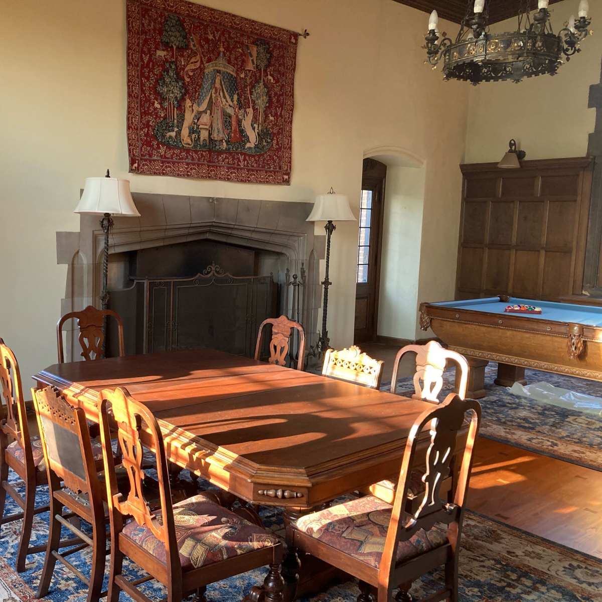A well-furnished dining room with a wooden table surrounded by six chairs, a large tapestry hanging on the wall, and a pool table in the background.