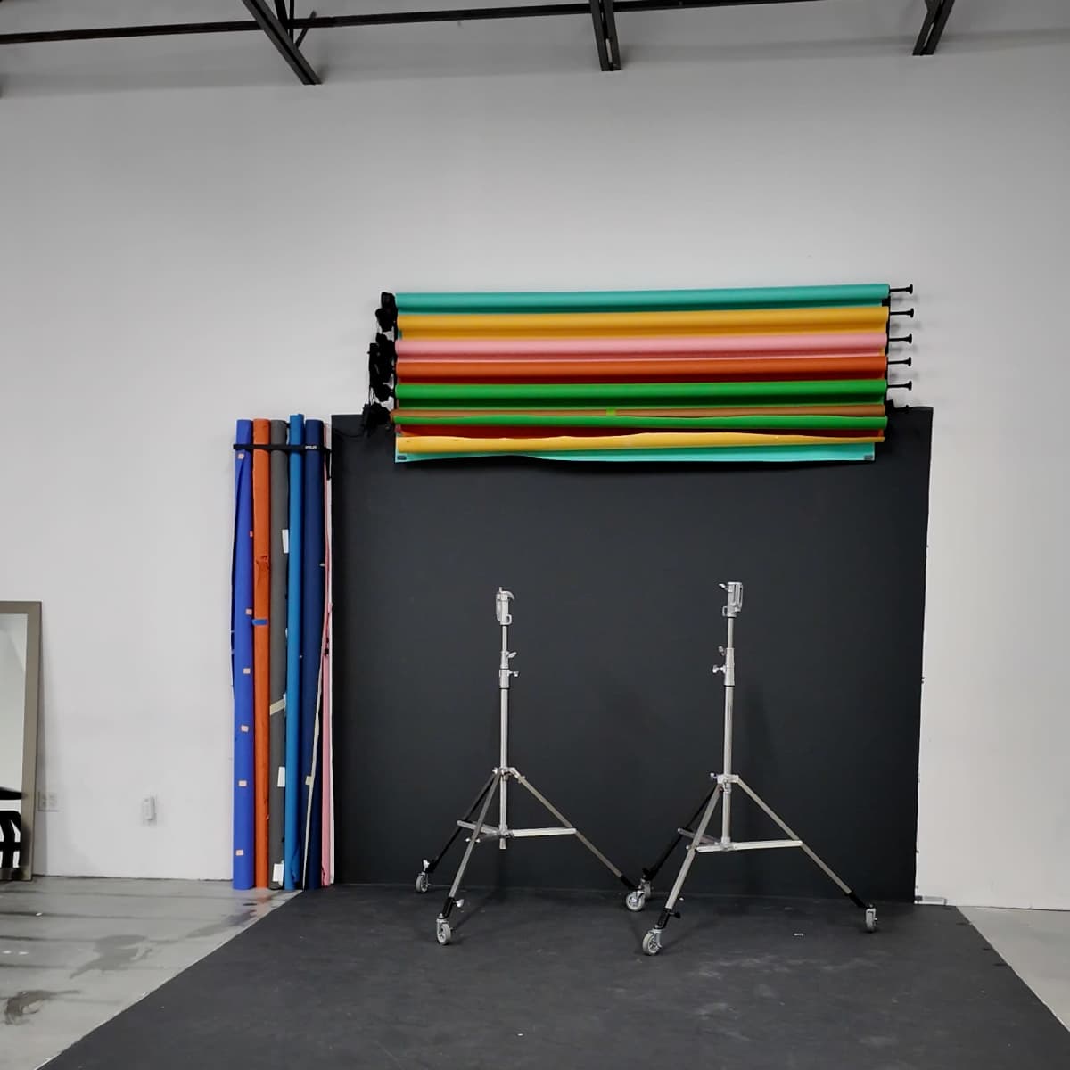 A photography studio setup with two lighting stands, a black backdrop, and colorful backdrop rolls stacked on a wall.