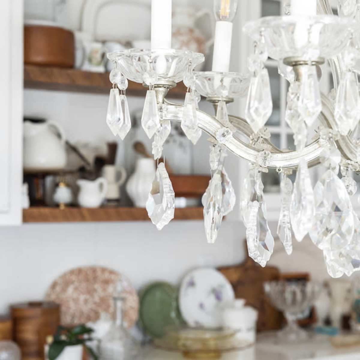 A crystal chandelier hangs above a shelf filled with various dishes and decorative items in a brightly lit kitchen.