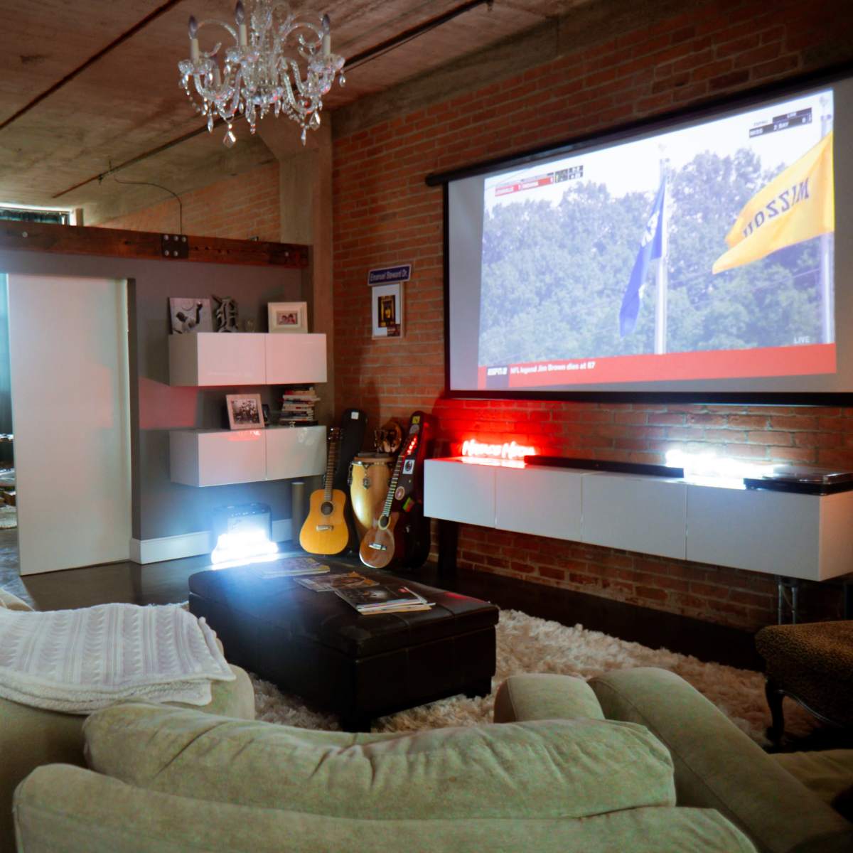 A modern living room with a large projector screen displaying a sports event, a sectional sofa, a coffee table, and guitars displayed alongside shelves on a brick wall.