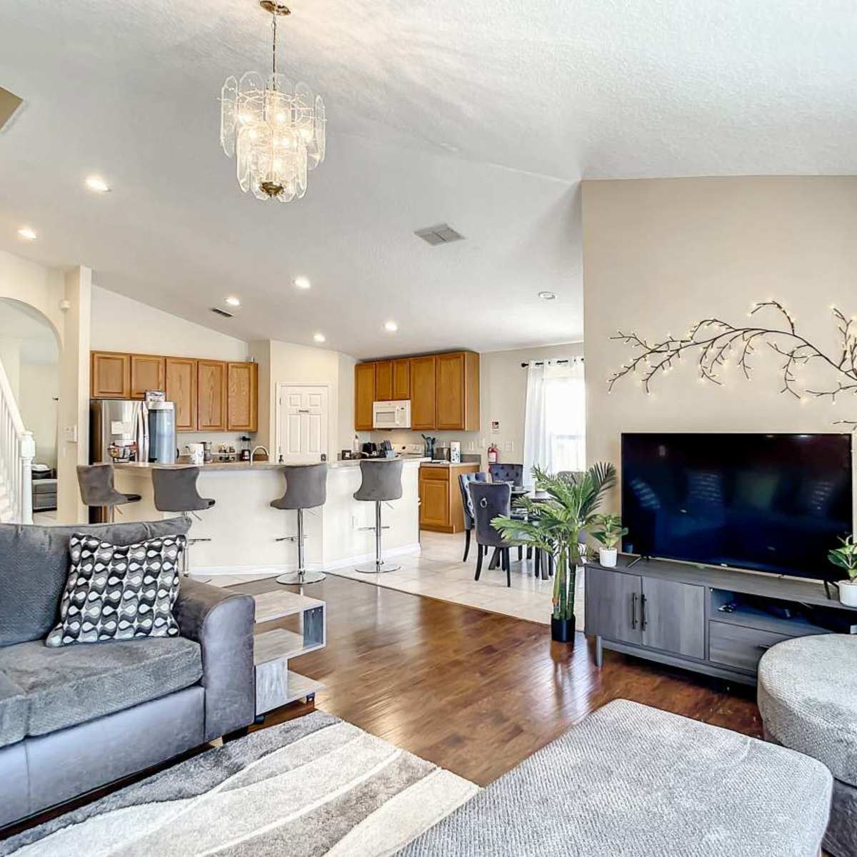 The living room features a large sectional sofa, a TV stand with a flat-screen television, and a modern kitchen visible in the background.