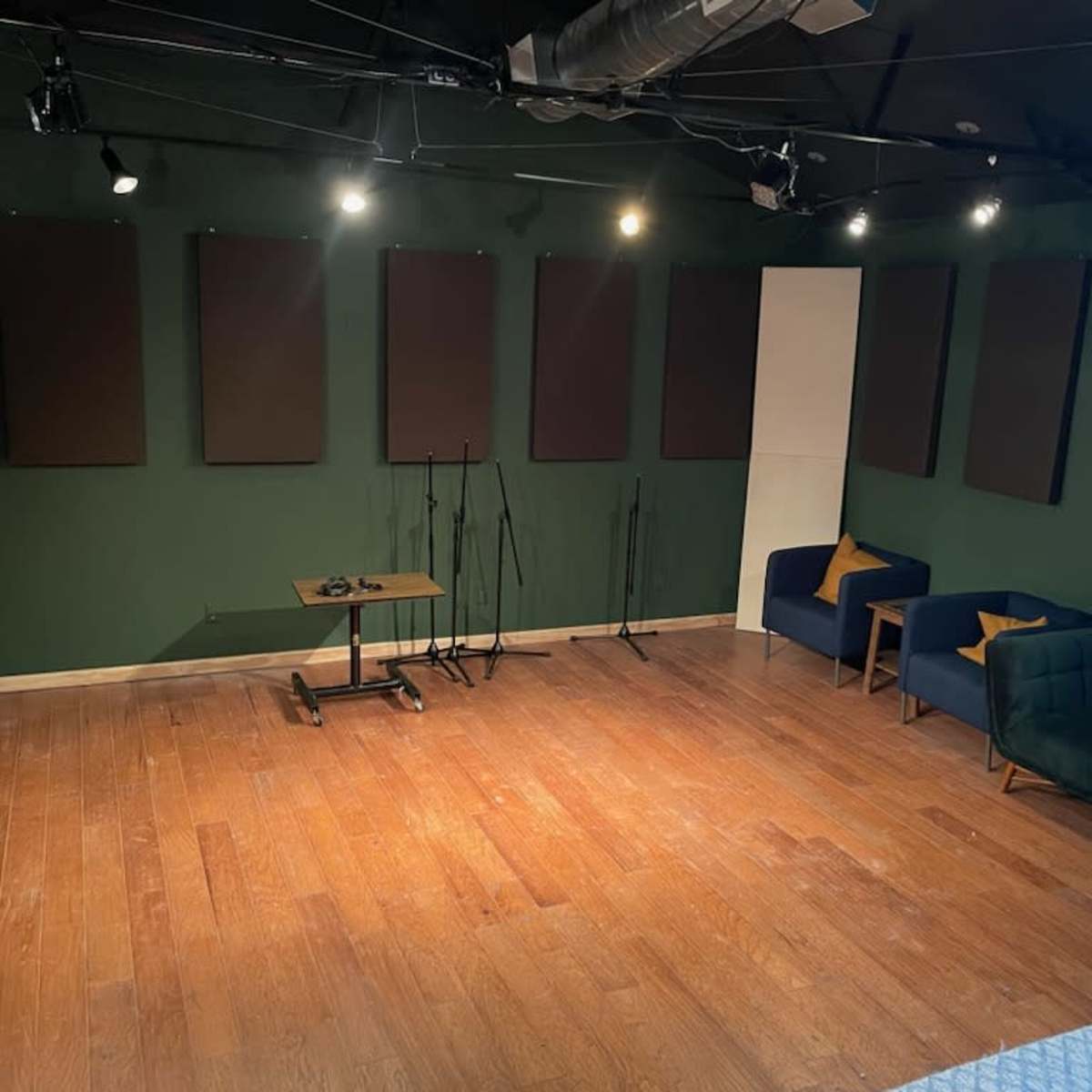 A music or recording studio with wooden flooring, green walls, multiple soundproof panels, and seating arrangements including two couches and a table.