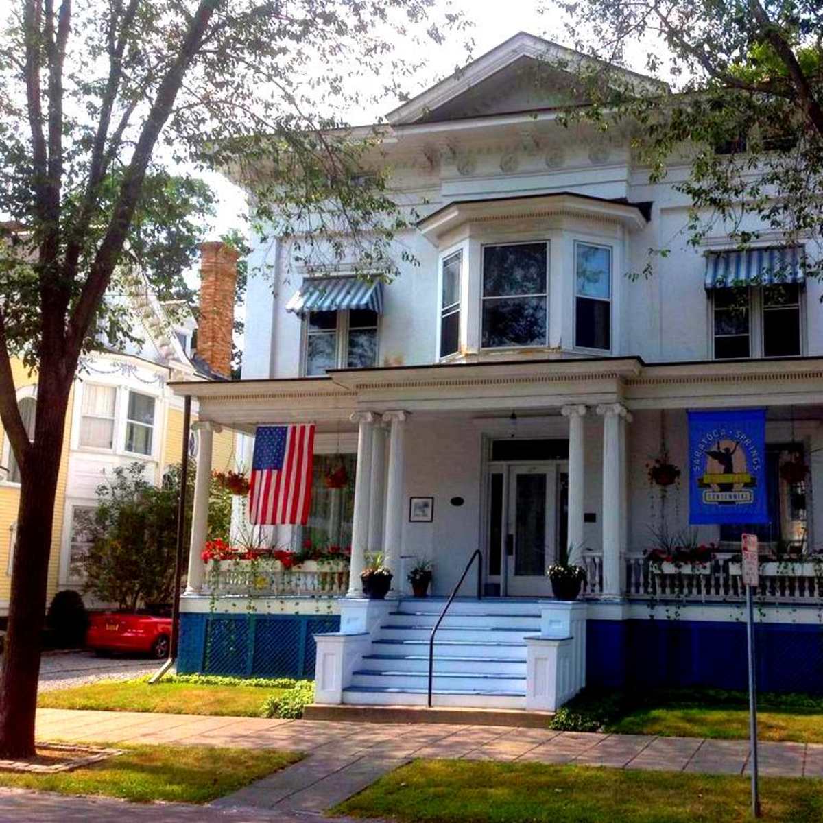 A large, two-story white house with a prominent porch, decorative elements, and an American flag hanging from the front.