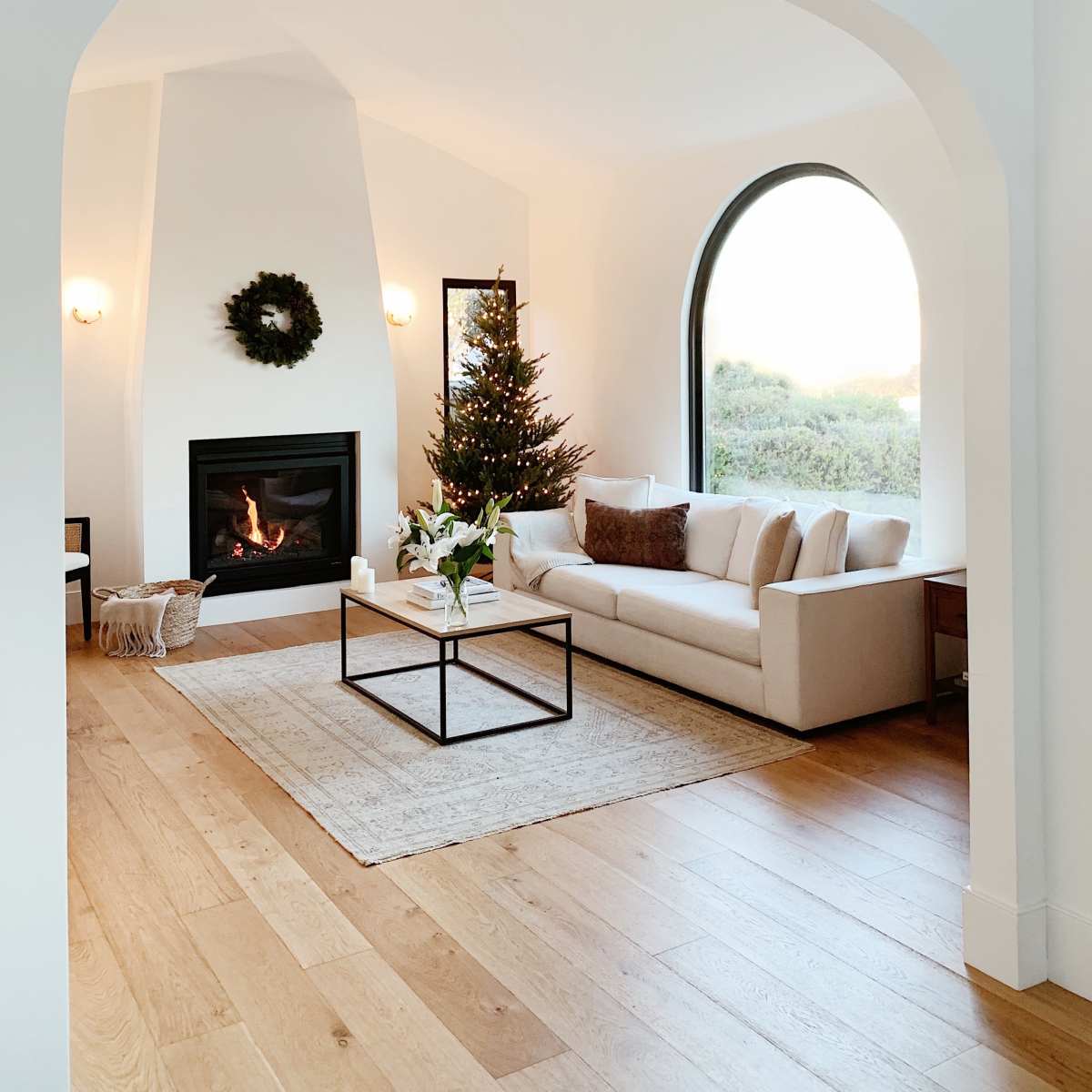A modern living room features a white sofa, a coffee table, a Christmas tree, and a fireplace, with an archway leading to a window that overlooks greenery.