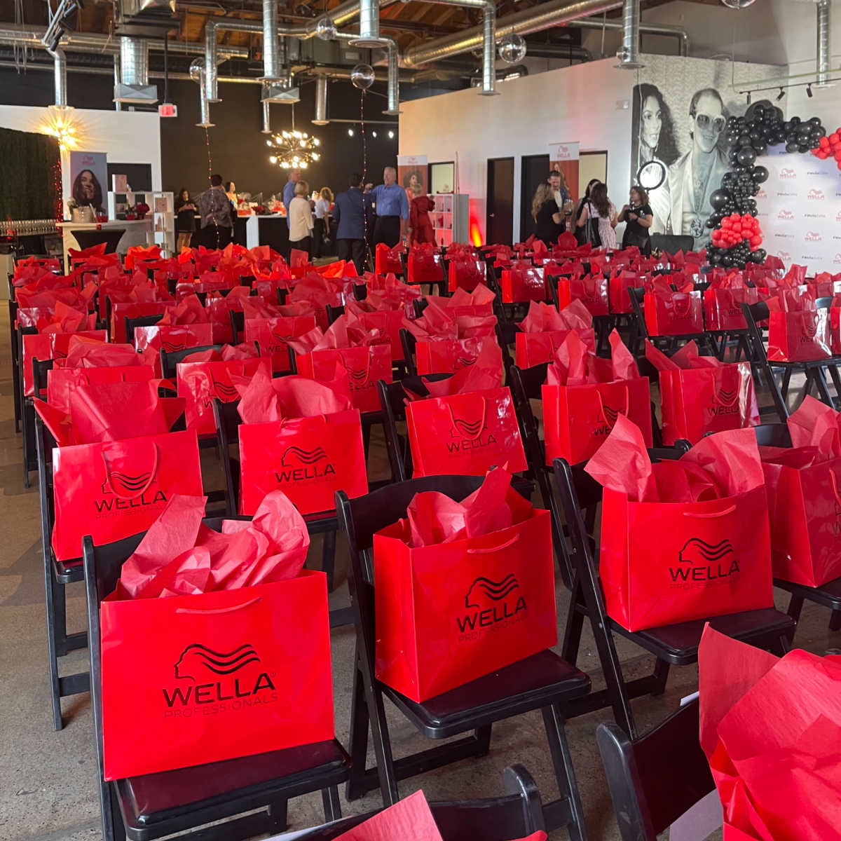 A venue with rows of black chairs arranged in front of tables, each chair featuring a red gift bag labeled "WELLA" filled with tissue paper.