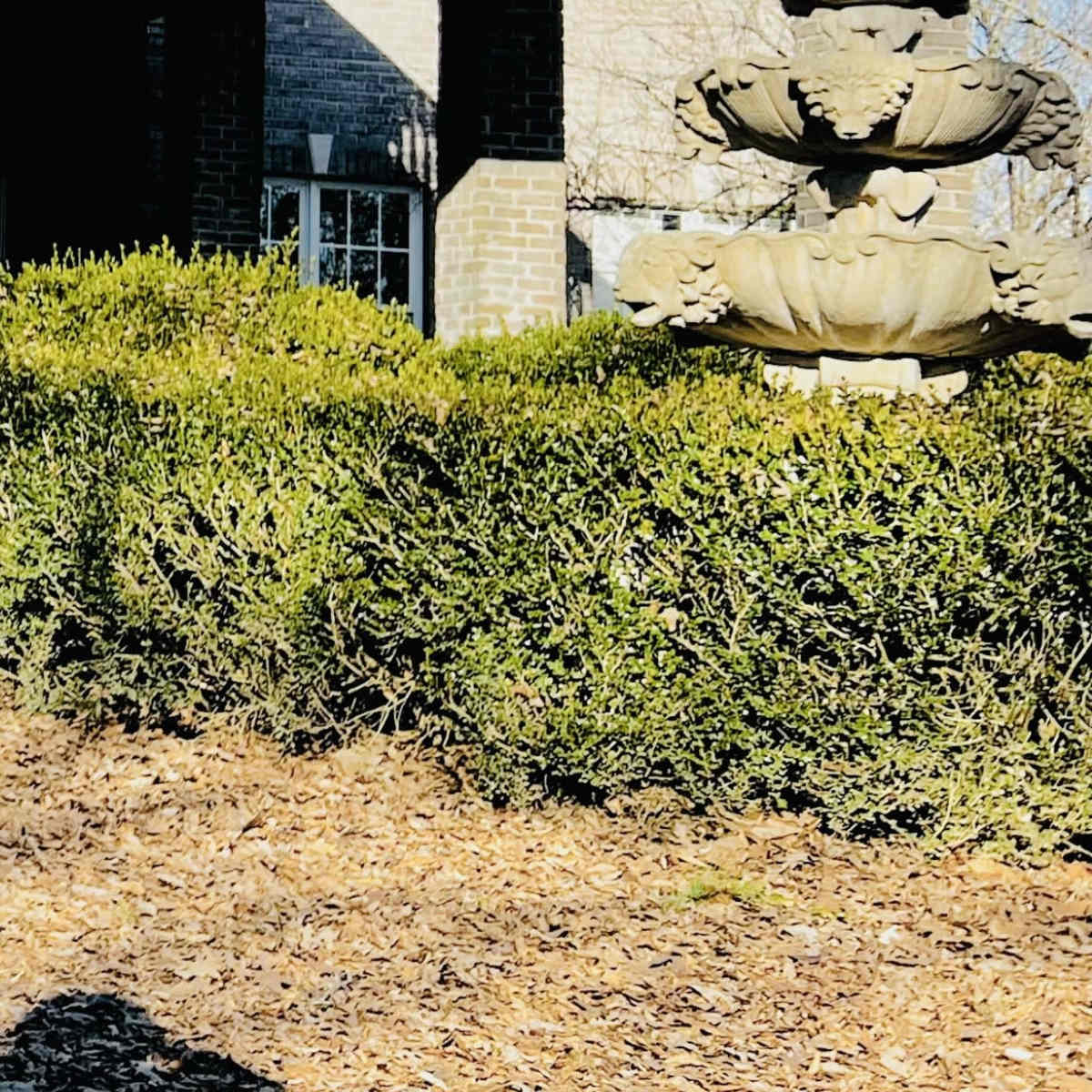 A stone fountain stands in a neatly trimmed garden under a decked balcony of a brick house.