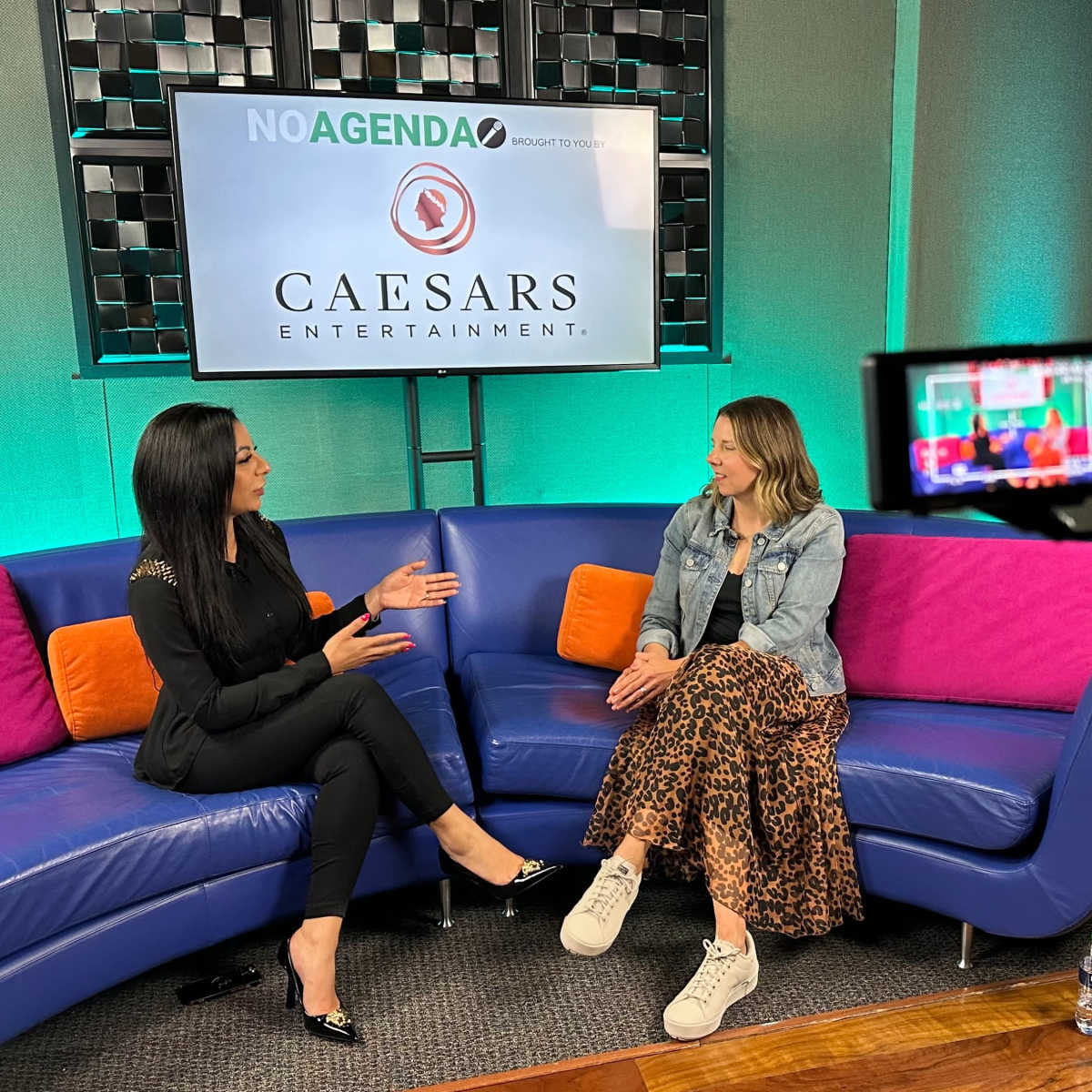 Two women sit on a colorful couch in a studio, engaged in conversation, with a camera and a logo for Caesars Entertainment visible in the background.