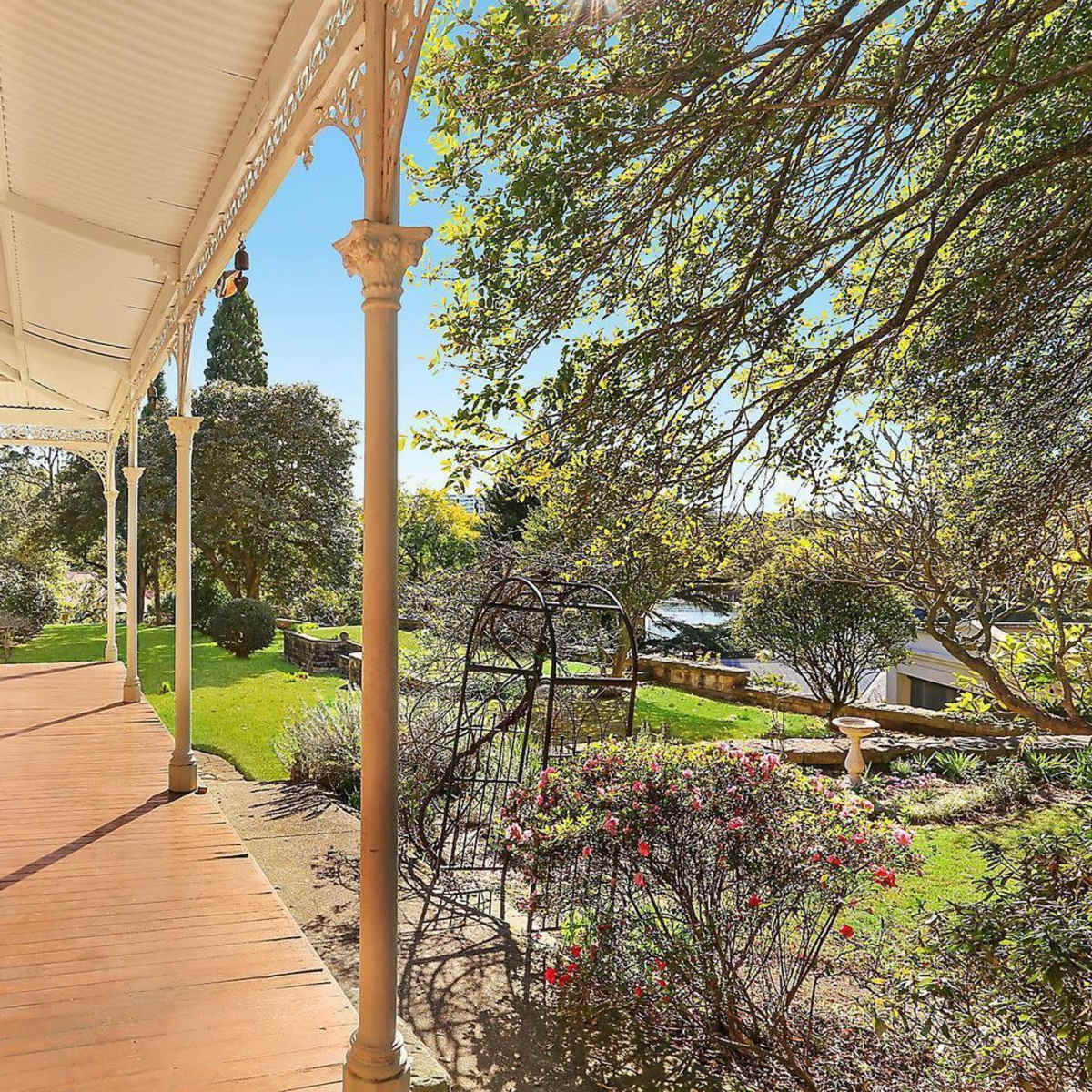 A porch with wooden flooring, supported by decorative columns, overlooking a garden with greenery and a stone pathway.