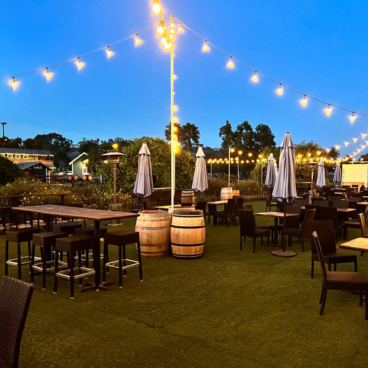 The scene shows a spacious outdoor dining area with tables and chairs surrounded by string lights and large umbrellas, set against a twilight sky.