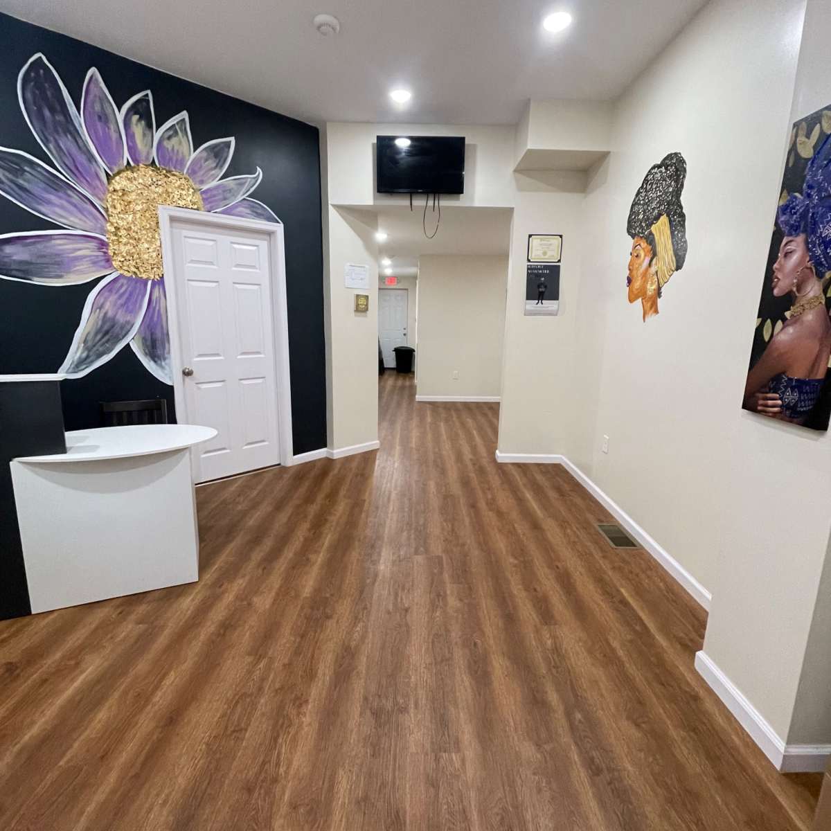 A sleek interior hallway with wooden flooring, a decorative wall art featuring a flower and portraits of women, and a small reception area on the left.