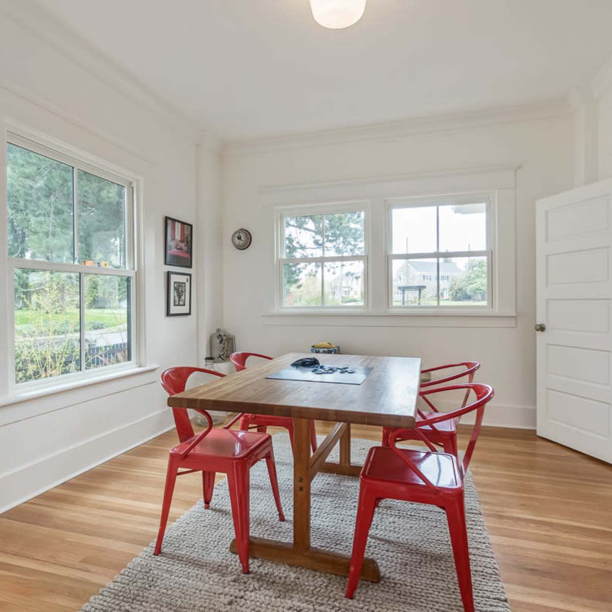 A modern dining room features a wooden table surrounded by four red chairs, with large windows allowing natural light to illuminate the space.