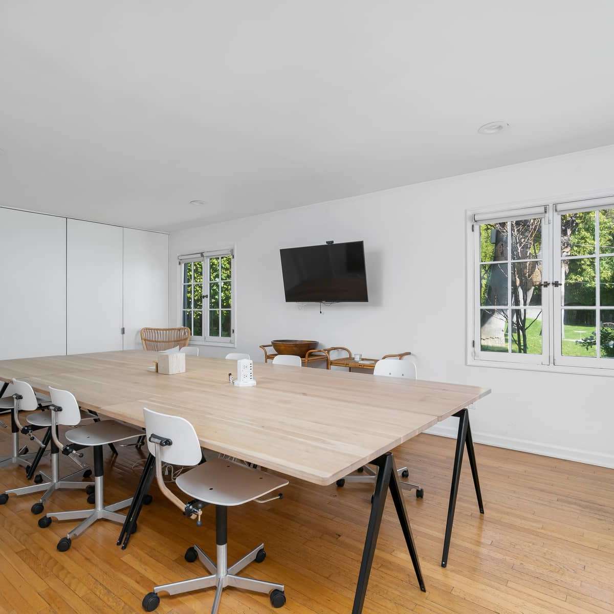 A large, simple conference table is surrounded by chairs in a bright, airy meeting room with windows and a wall-mounted television.