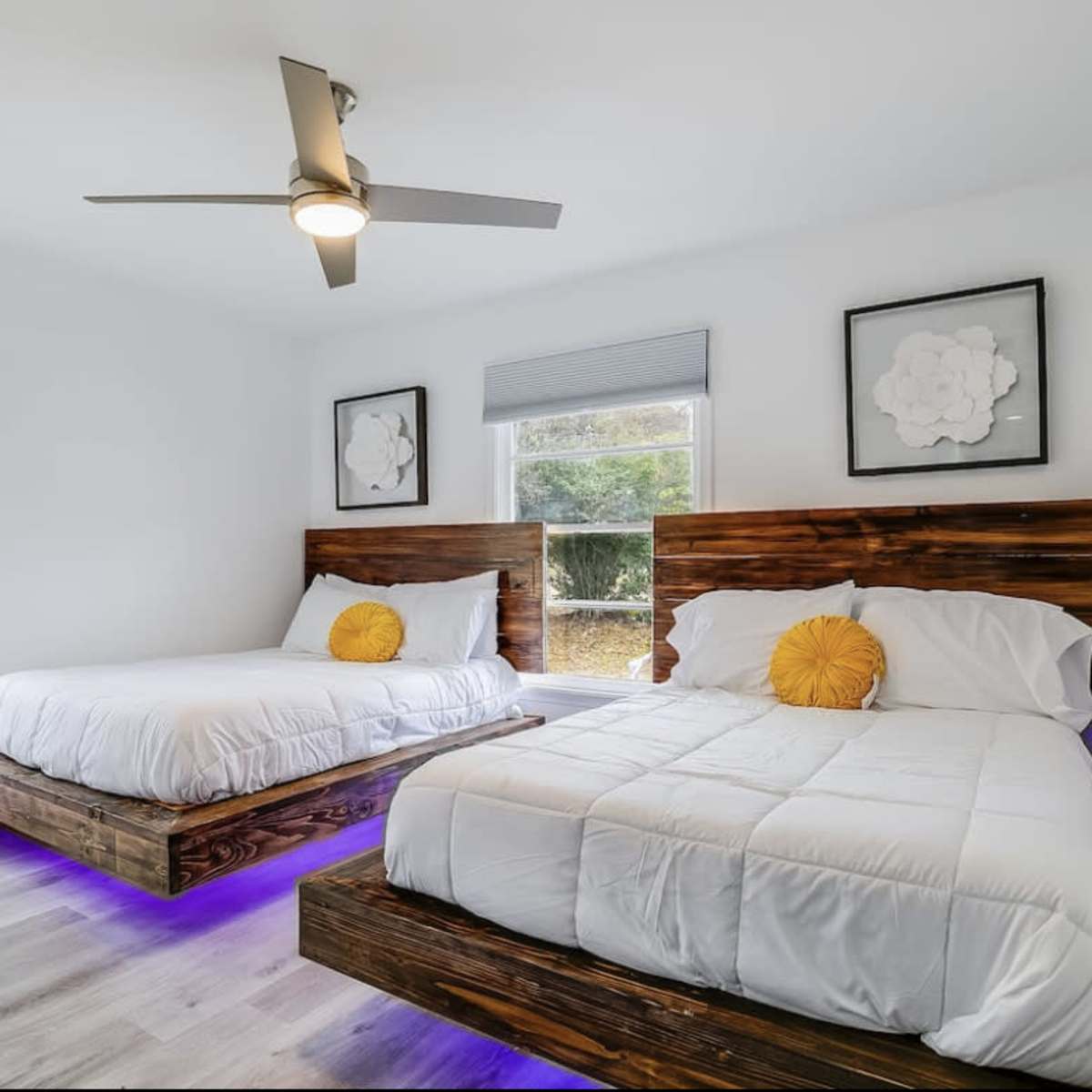 A bright bedroom featuring two wooden platform beds with white bedding and yellow accent pillows, alongside large windows letting in natural light.