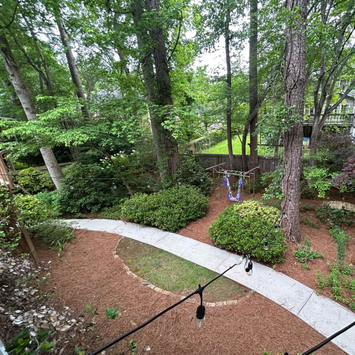 A winding concrete path leads through a lush, wooded garden featuring several trees and shrubs.