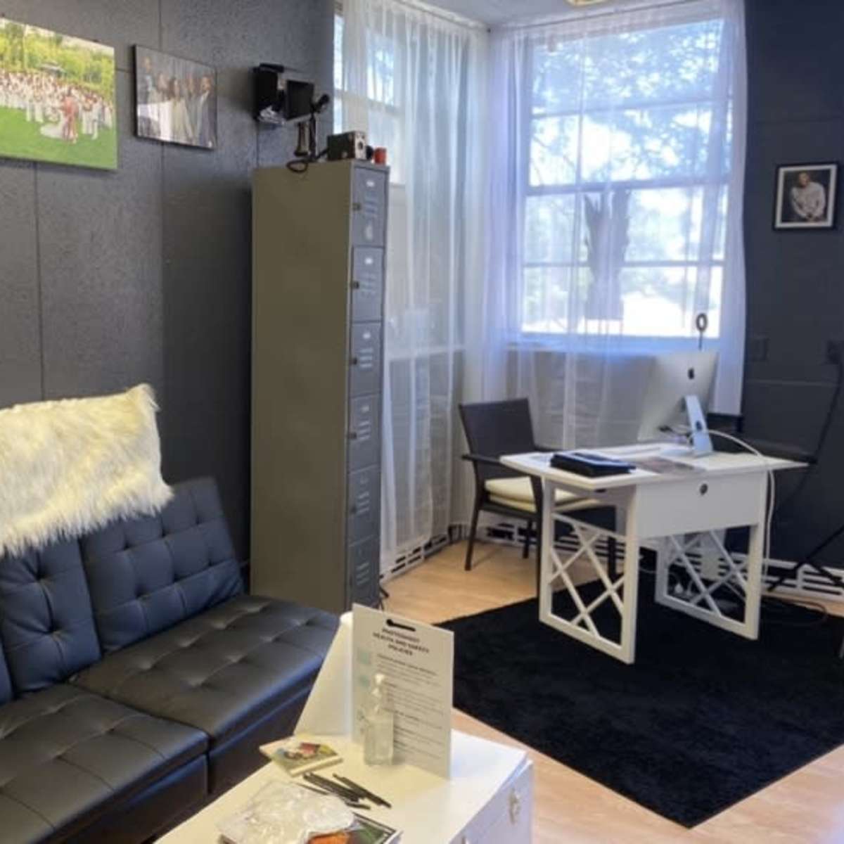 A modern office space featuring a black leather couch with a white fur throw, a desk with a computer, and large windows letting in natural light.