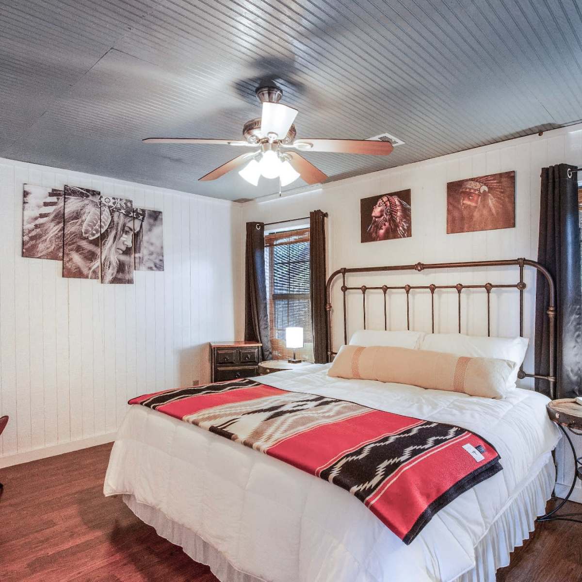 A neatly arranged bedroom features a metal bed frame with a white comforter, decorative pillows, and wooden flooring, alongside large windows with blinds.