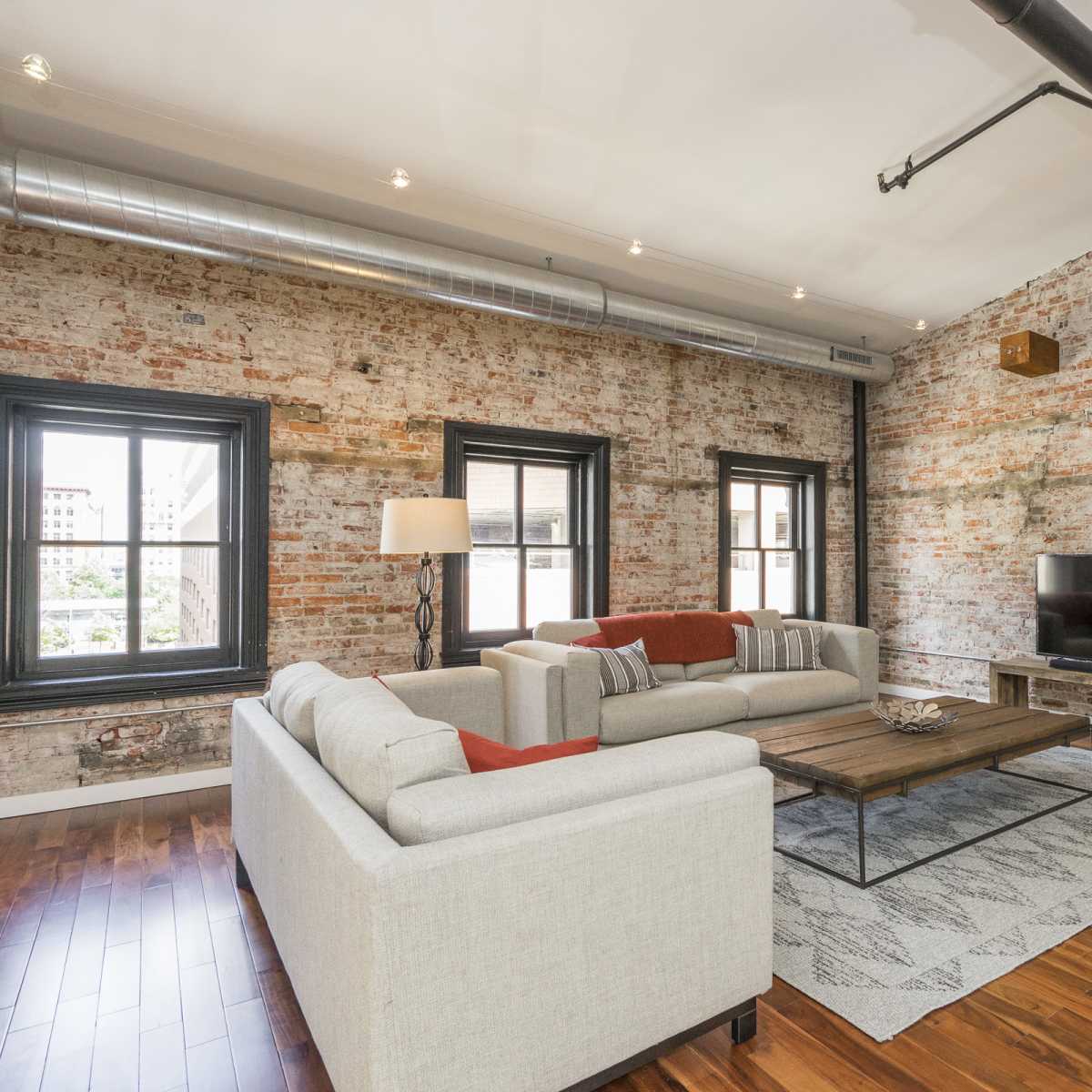 A modern living room with exposed brick walls, hardwood floors, a sectional sofa, a coffee table, and a television mounted on the wall.