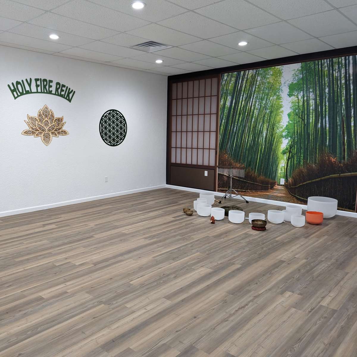 An interior of a meditation or wellness studio featuring wood flooring, a large mural of a bamboo forest, decorative wall elements, and various sound bowls arranged on the floor.