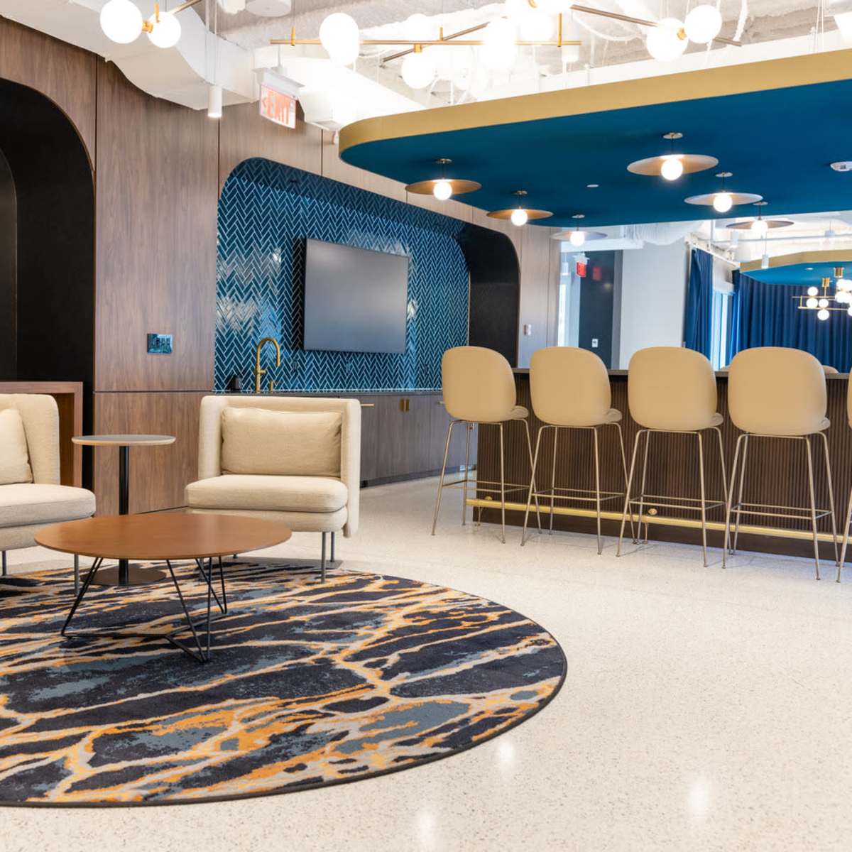 A modern lounge area features light-colored seating arranged around a circular coffee table, with a bar in the background and decorative lighting overhead.