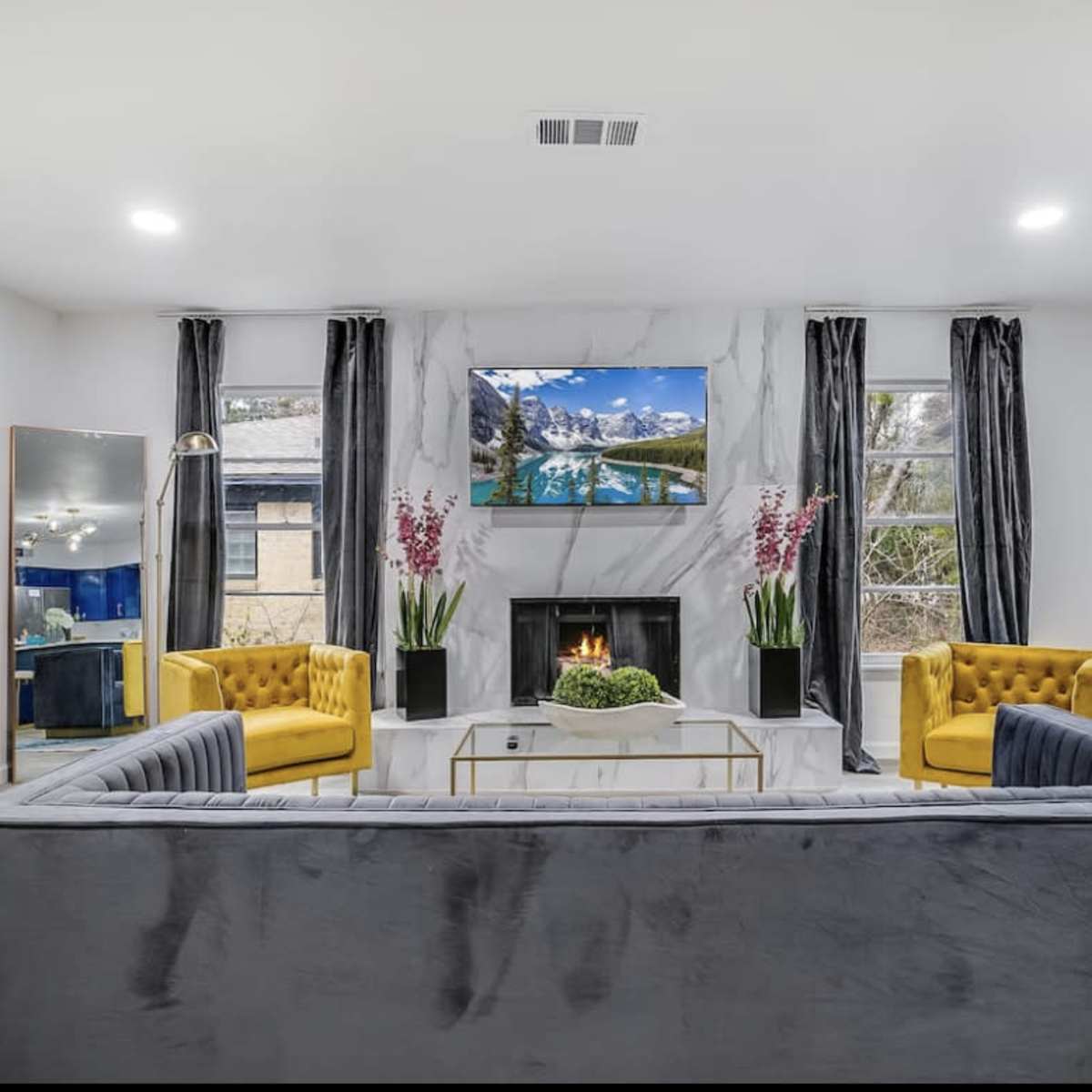 A modern living room features a gray sofa, two yellow chairs, a marble fireplace, and a wall-mounted TV above the mantel.