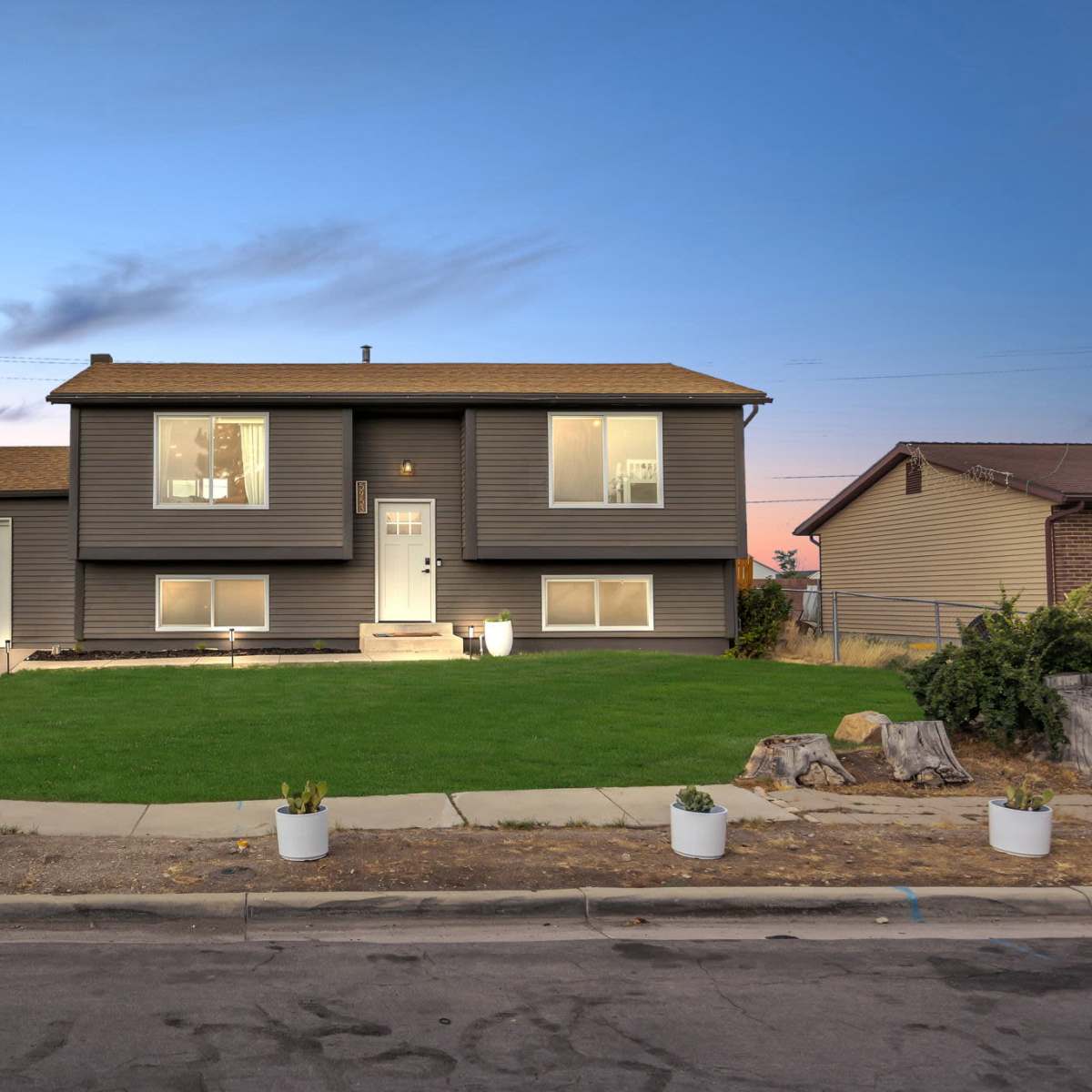 A two-story gray house with a front lawn and a driveway is situated on a suburban street at dusk.