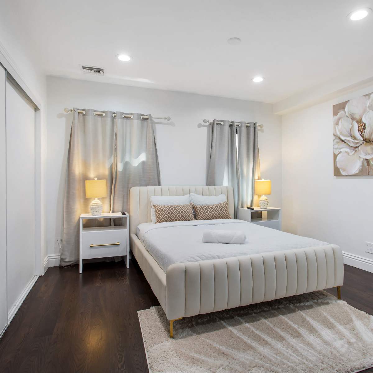 A modern bedroom features a white upholstered bed frame, two bedside tables with lamps, and floral artwork on the wall, complemented by gray curtains.