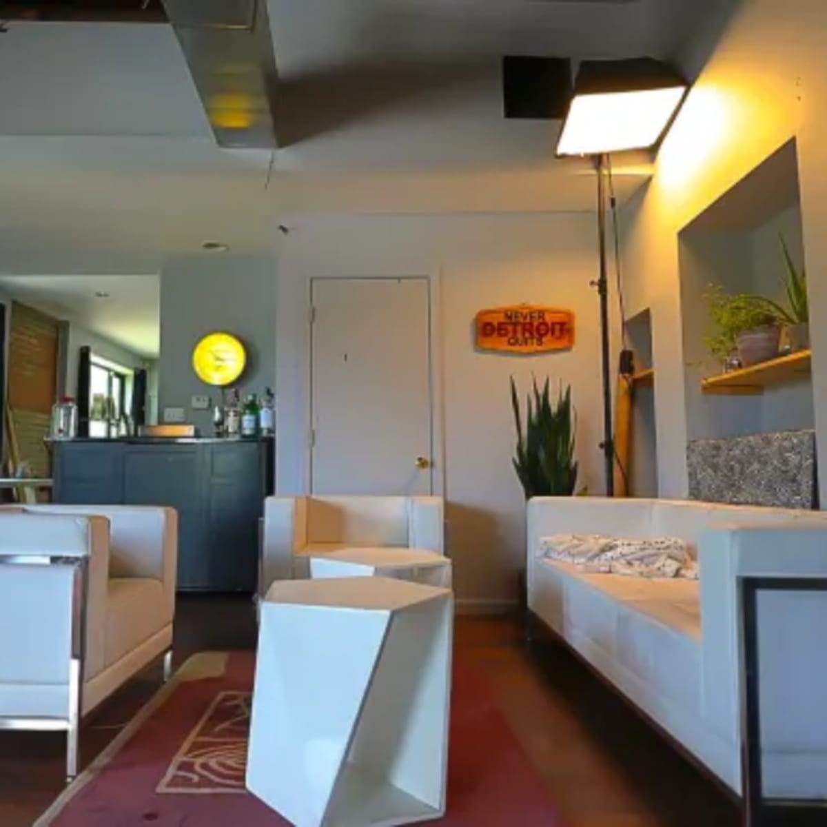 A modern lounge area with white furniture, a bar counter stocked with bottles, and a triangular coffee table.