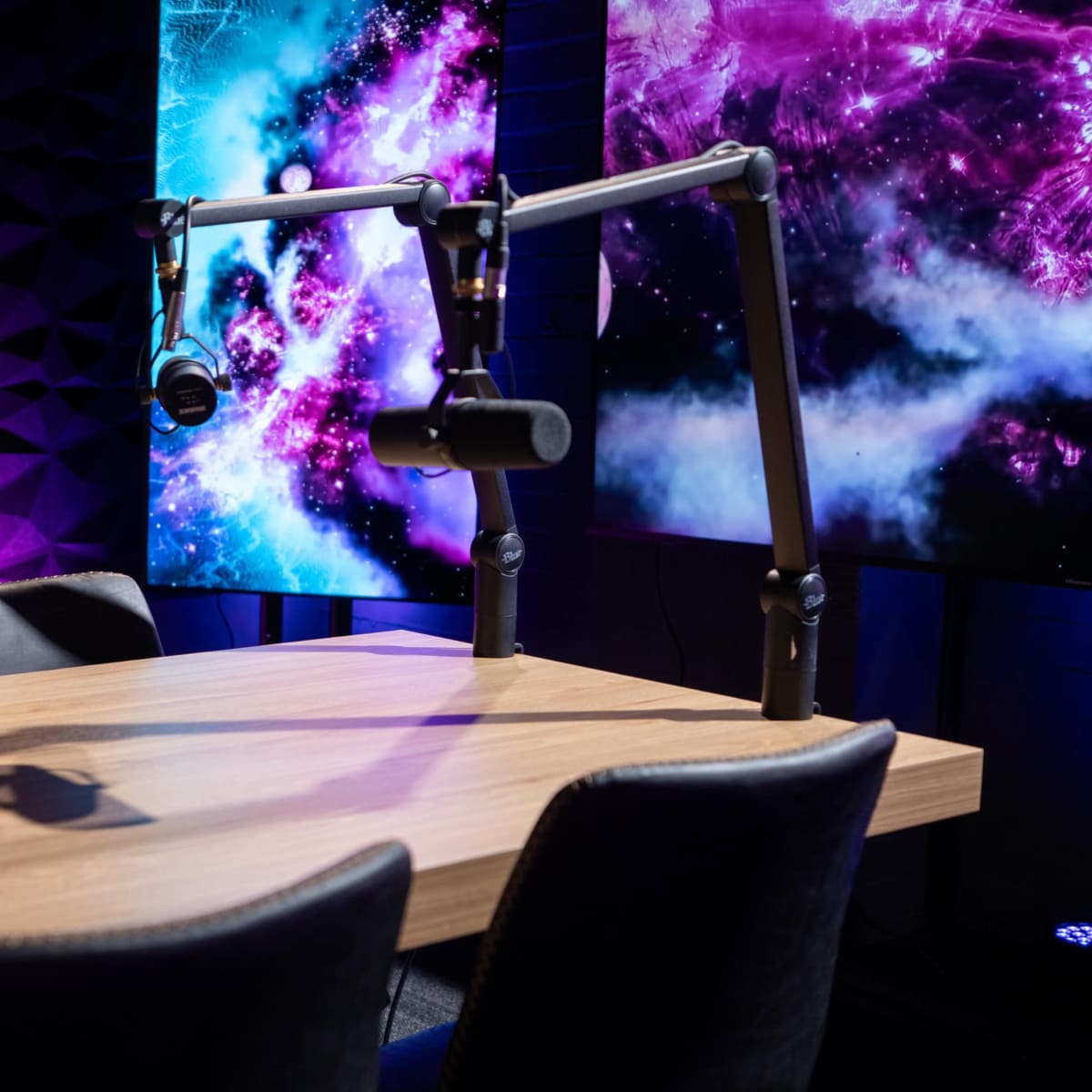 A podcast studio features a wooden table with black chairs and microphone stands, set against a colorful backdrop of purple and blue graphics.