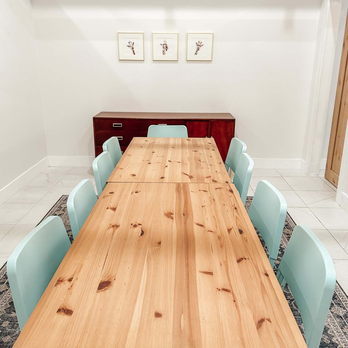 A wooden dining table with eight light blue chairs is set against a plain wall featuring three framed artworks above a red storage unit.