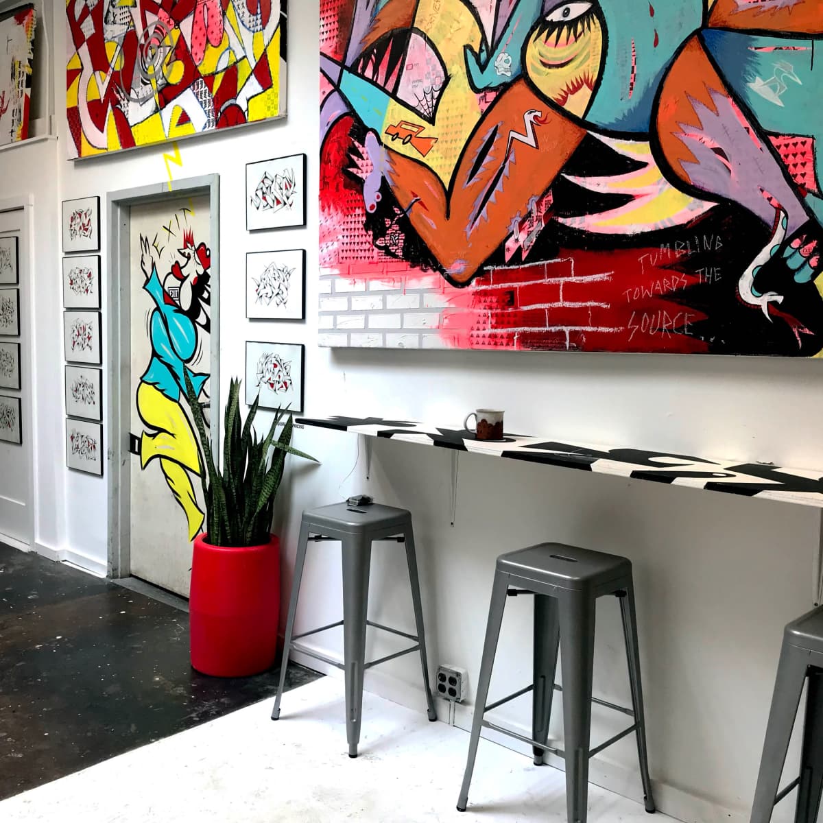 A modern art space featuring brightly colored murals on the walls, a red planter, and metal stools arranged near a sleek counter.