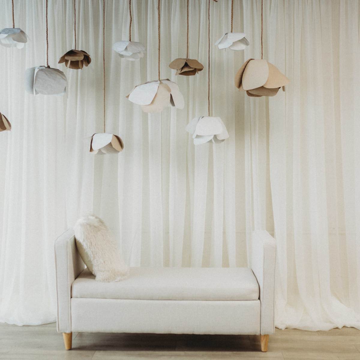 A minimalistic room with a cream-colored chaise lounge beneath a backdrop of flowing white curtains and hanging paper flower decorations.