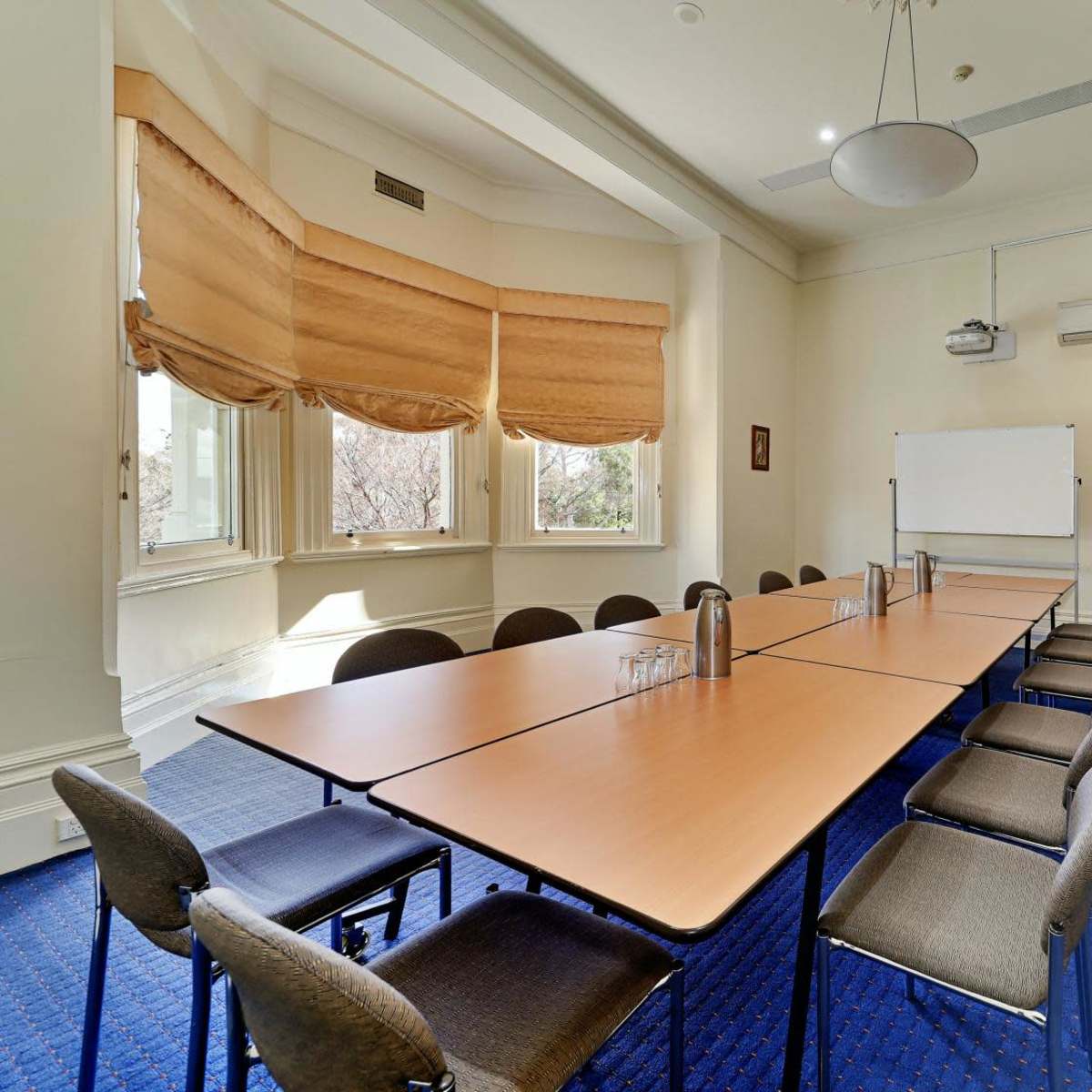 A rectangular meeting room features a long table with several chairs, large windows with Roman shades, and a whiteboard on the wall.