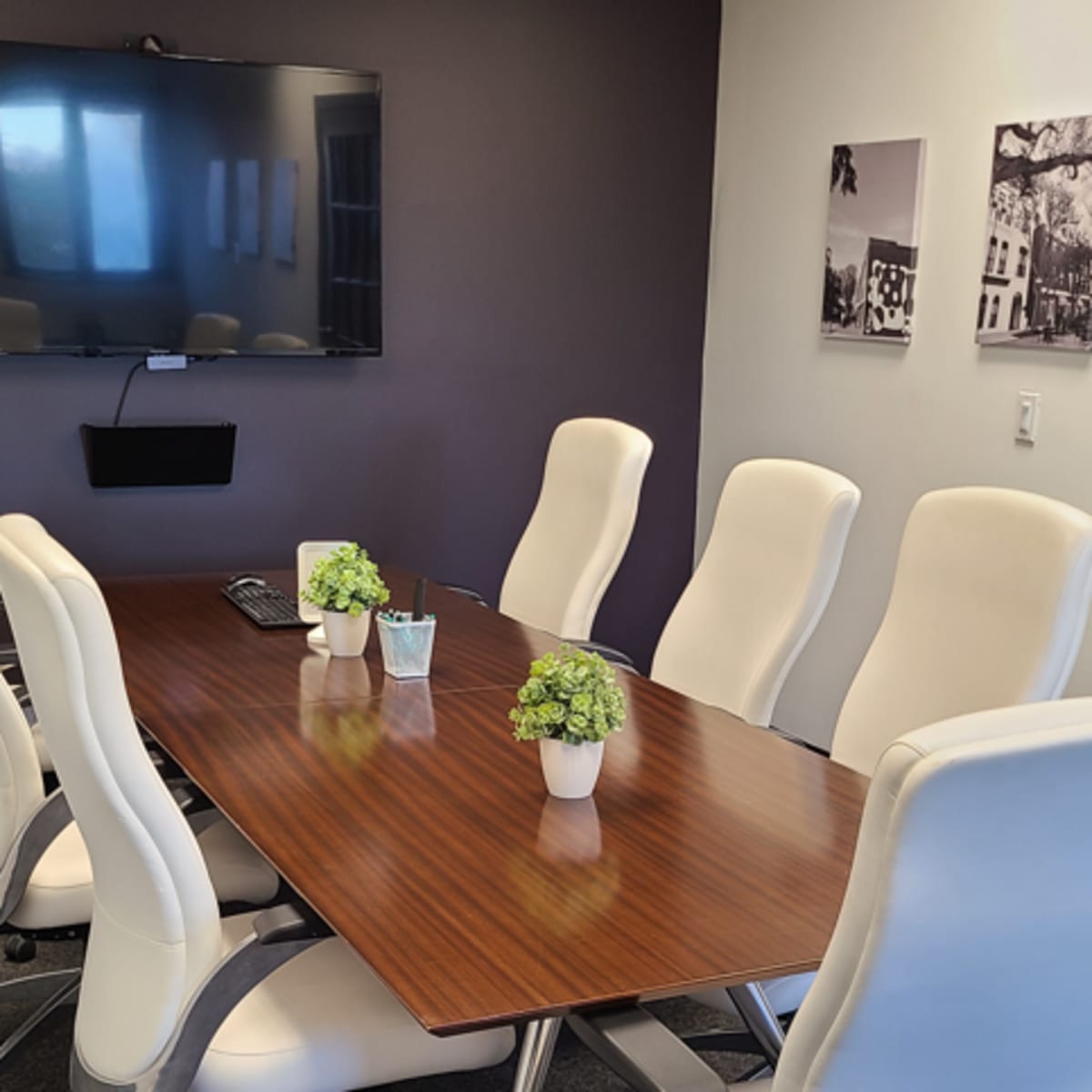 A conference room features a long wooden table surrounded by white chairs, with a large screen on one wall and framed black-and-white photographs on another.