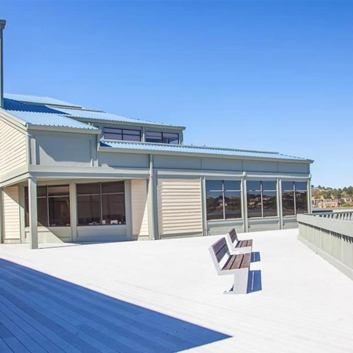 A large wooden deck with two benches, overlooking a body of water and a multi-level building with large windows.