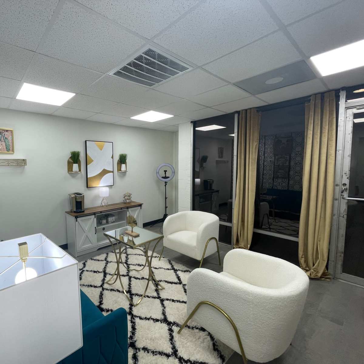 A modern waiting room with two white chairs, a glass coffee table, a blue accent sofa, and decorative wall art.