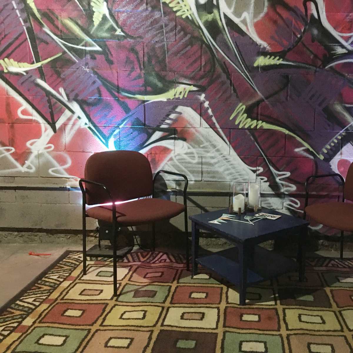 Two red chairs are positioned around a blue table on a patterned rug, with a colorful graffiti mural on the wall behind them.