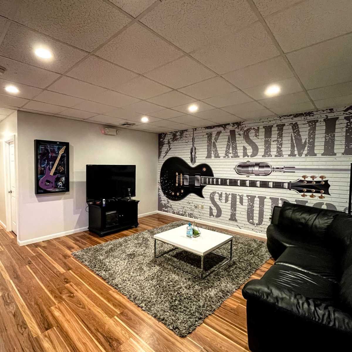 A living room with a black leather couch, a TV stand, and a wall featuring a guitar-themed mural, alongside a hallway leading to other rooms.