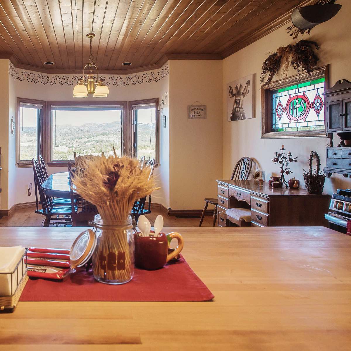 A brightly lit kitchen with wooden beams, featuring a central table adorned with a jar of dried flowers and red napkins, overlooks a scenic view through large windows.