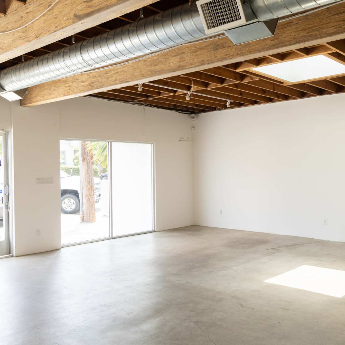 An empty room with concrete flooring, exposed wooden beams on the ceiling, and a large sliding glass door leading outside.