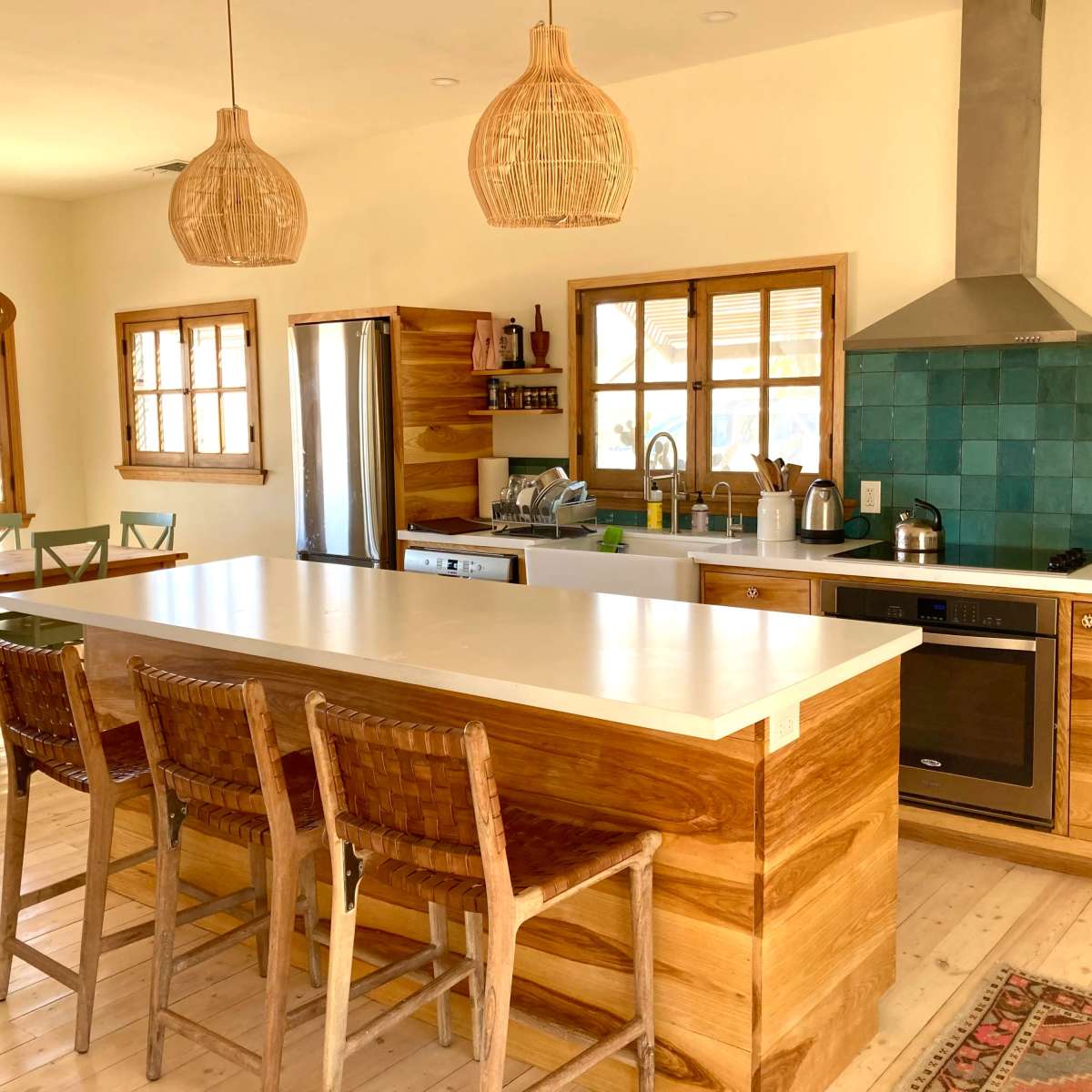 A modern kitchen features wooden cabinetry, a large island with seating, and a teal tile backsplash, with a dining area visible in the background.