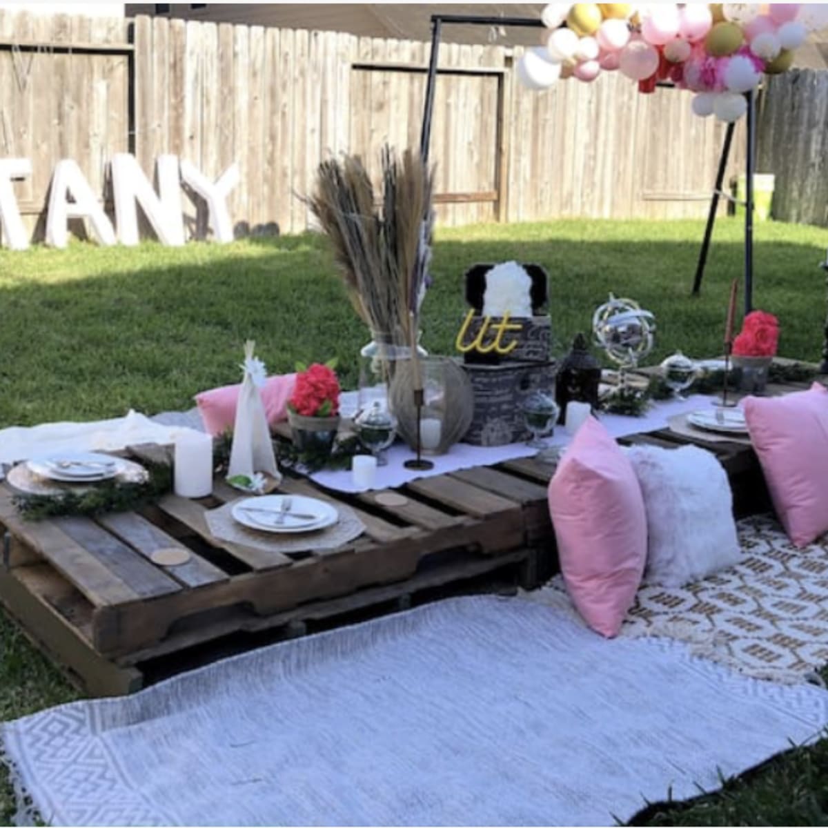 A decorated outdoor picnic setup on grass, featuring a low wooden pallet table adorned with plates, glasses, and decorative items, alongside large letters spelling "BRITTANY" and a floral arrangement overhead.