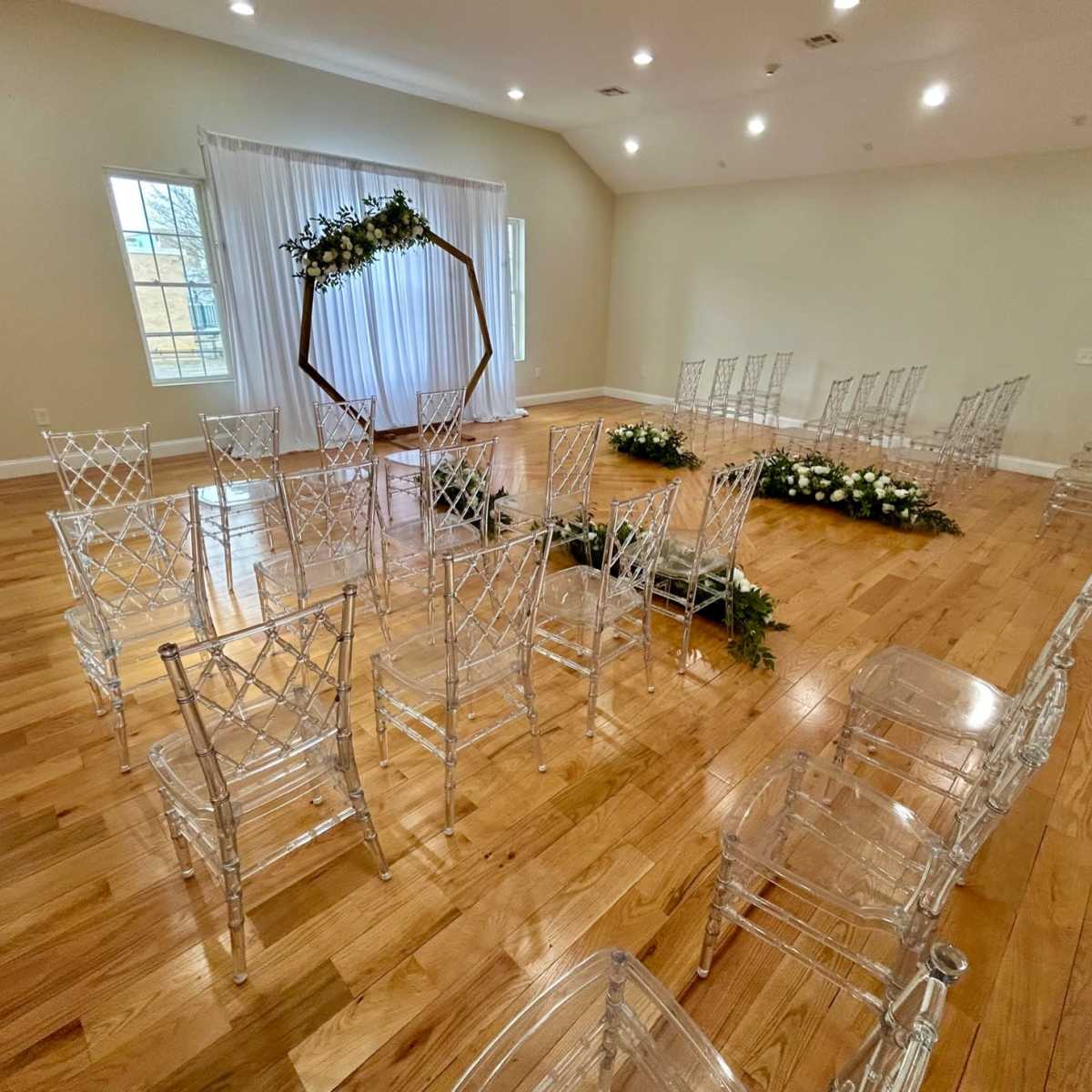 A spacious event room arranged with clear chairs in a semicircular formation around a floral-decorated archway and white drapery.