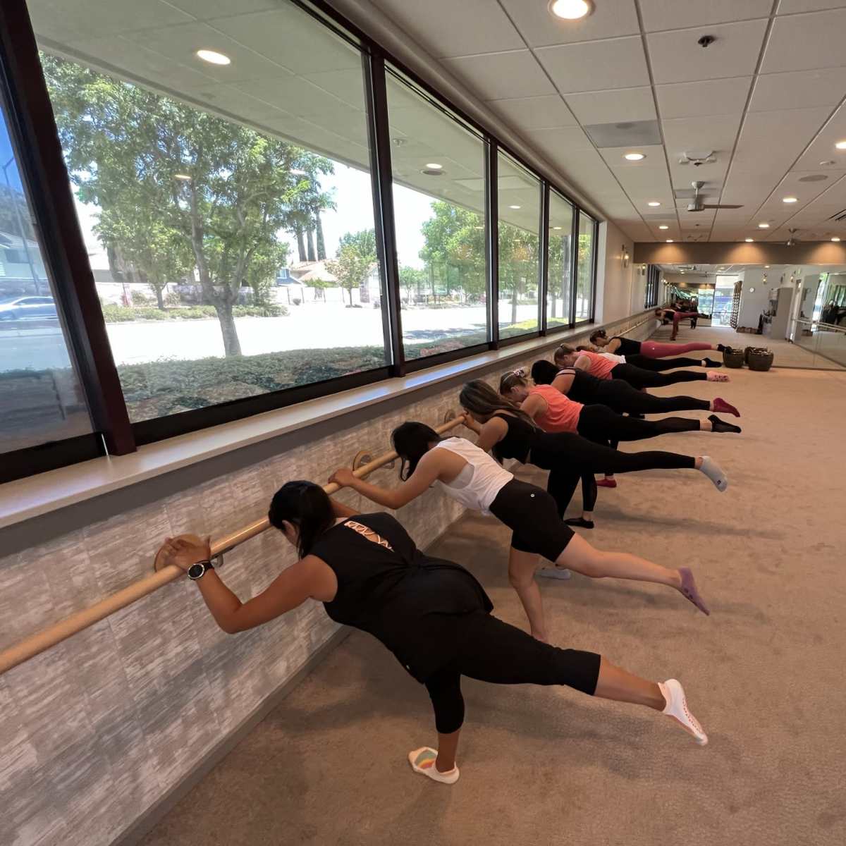 A group of individuals is performing barre exercises along a wooden ballet bar in a spacious studio with large windows.